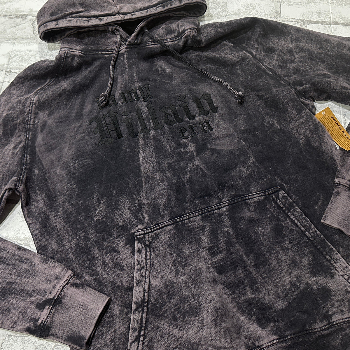 Black mineral-wash pullover hoodie layed flat, featuring black gothic lettering that reads ‘in my Villain era.