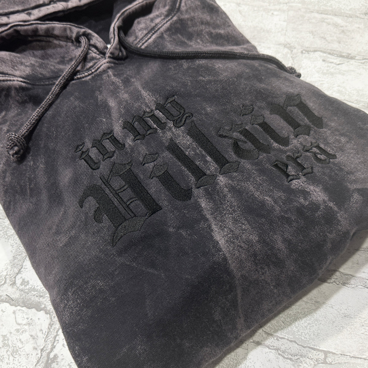 Black mineral-wash pullover hoodie folded flat, featuring black gothic lettering that reads ‘in my Villain era.