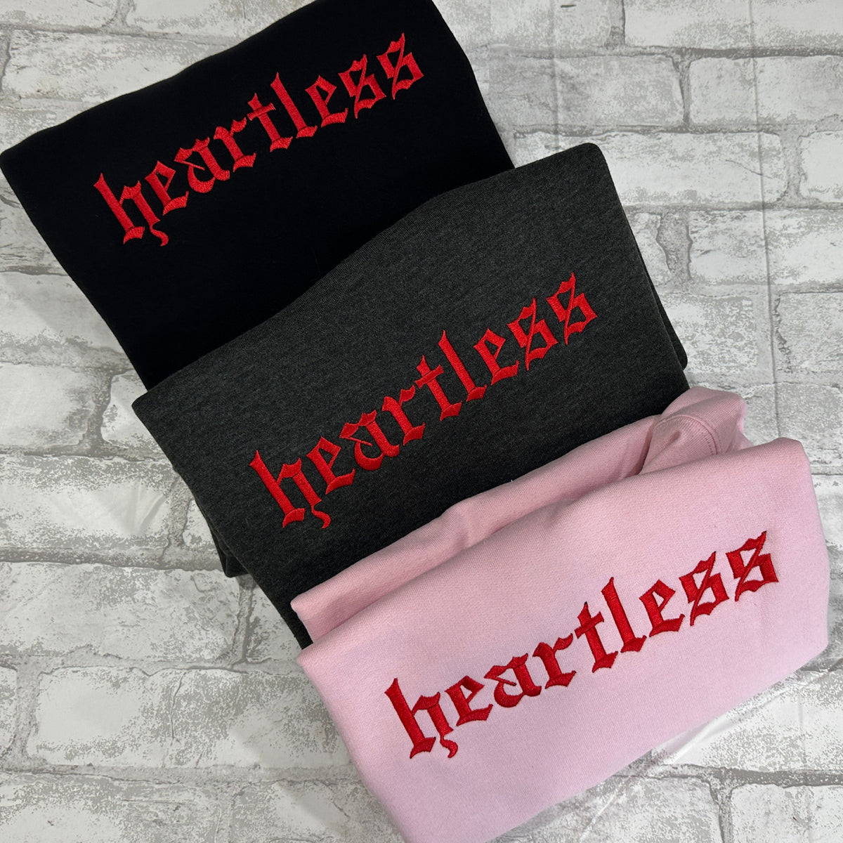 Three crewneck sweatshirts, one black, one dark grey, and one pink, laid flat with the word 'heartless' embroidered in red on the front.