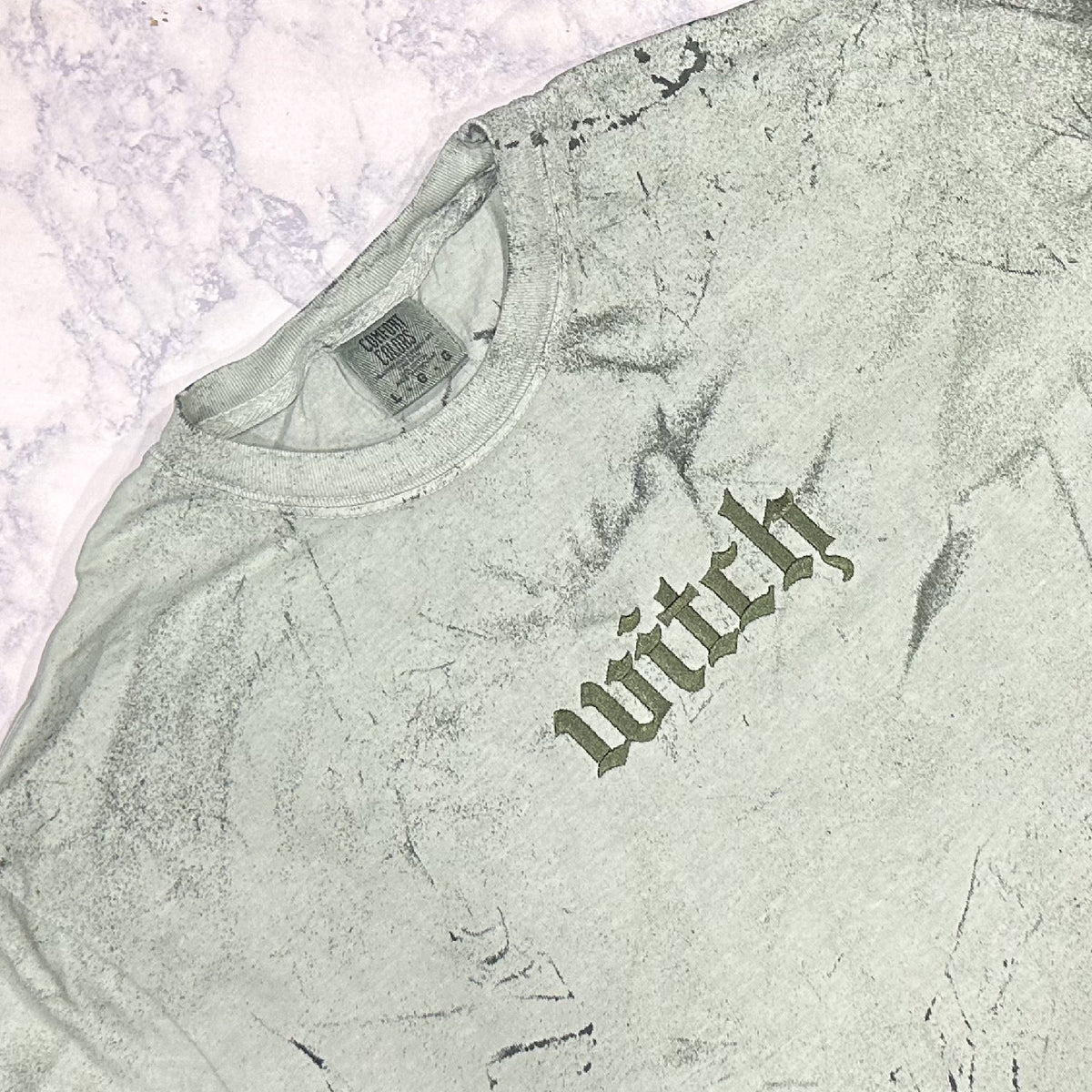 Light forest green mineral-washed T-shirt laid flat, featuring small tonal embroidered text reading “witch” on the chest.