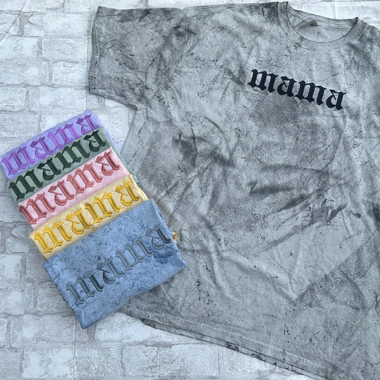 Flat lay of a mineral-wash gray T-shirt with black embroidered text reading “mama,” shown with folded color options embroidered with the same design.