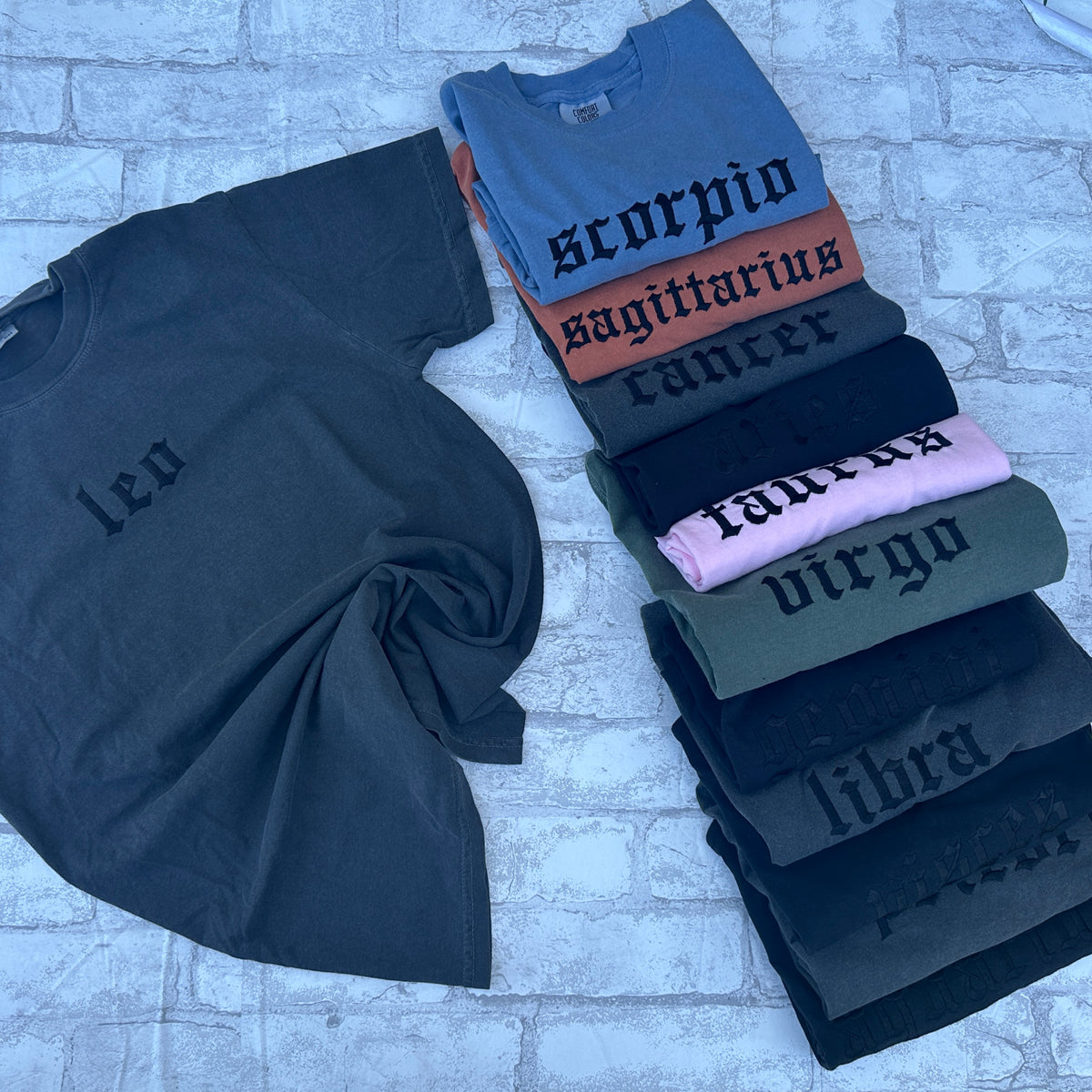 A collection of unisex fit t-shirts with embroidered zodiac signs in various colors displayed on a textured surface.