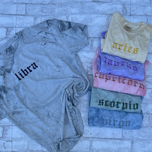A collection of embroidered t-shirts with various zodiac signs displayed on a stone surface. The main shirt is gray with the zodiac Libra embroidered on the front, and others have different signs like Aries, Capricorn, Scorpio, and Pisces.