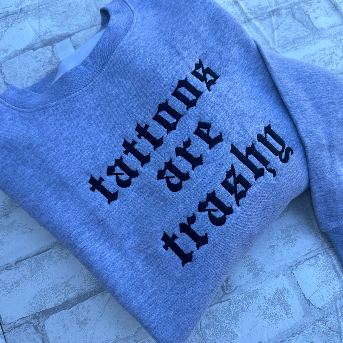 heather gray crewneck sweatshirt featuring black embroidered “tattoos are trashy” lettering in lowercase Old English font, stacked across three lines on the chest.