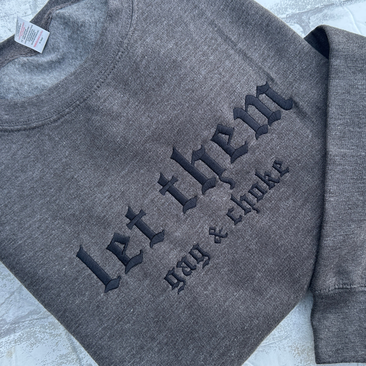 Dark Heather gray crewneck sweatshirt featuring black embroidered gothic text across the chest reading “let them” with smaller text underneath reading “gag & choke,” shown flat on a white background.