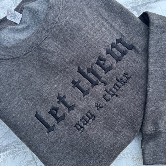Dark Heather gray crewneck sweatshirt featuring black embroidered gothic text across the chest reading “let them” with smaller text underneath reading “gag & choke,” shown flat on a white background.