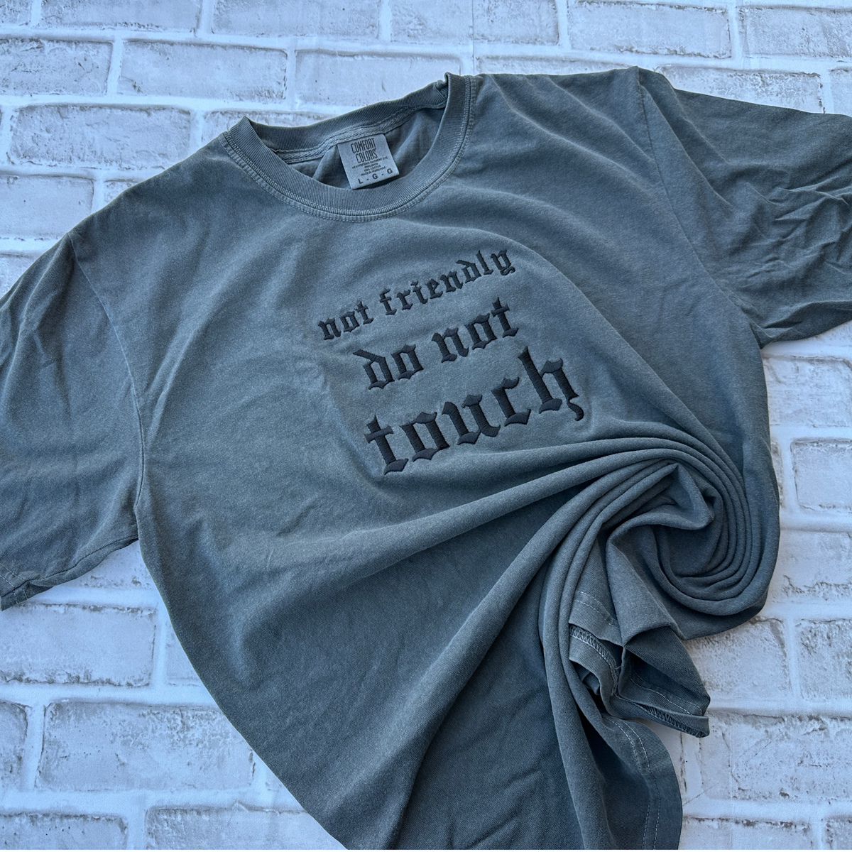 Pepper Comfort Colors short sleeve t-shirt featuring black Gothic-style embroidery reading “not friendly do not touch” across the center chest in three lines.