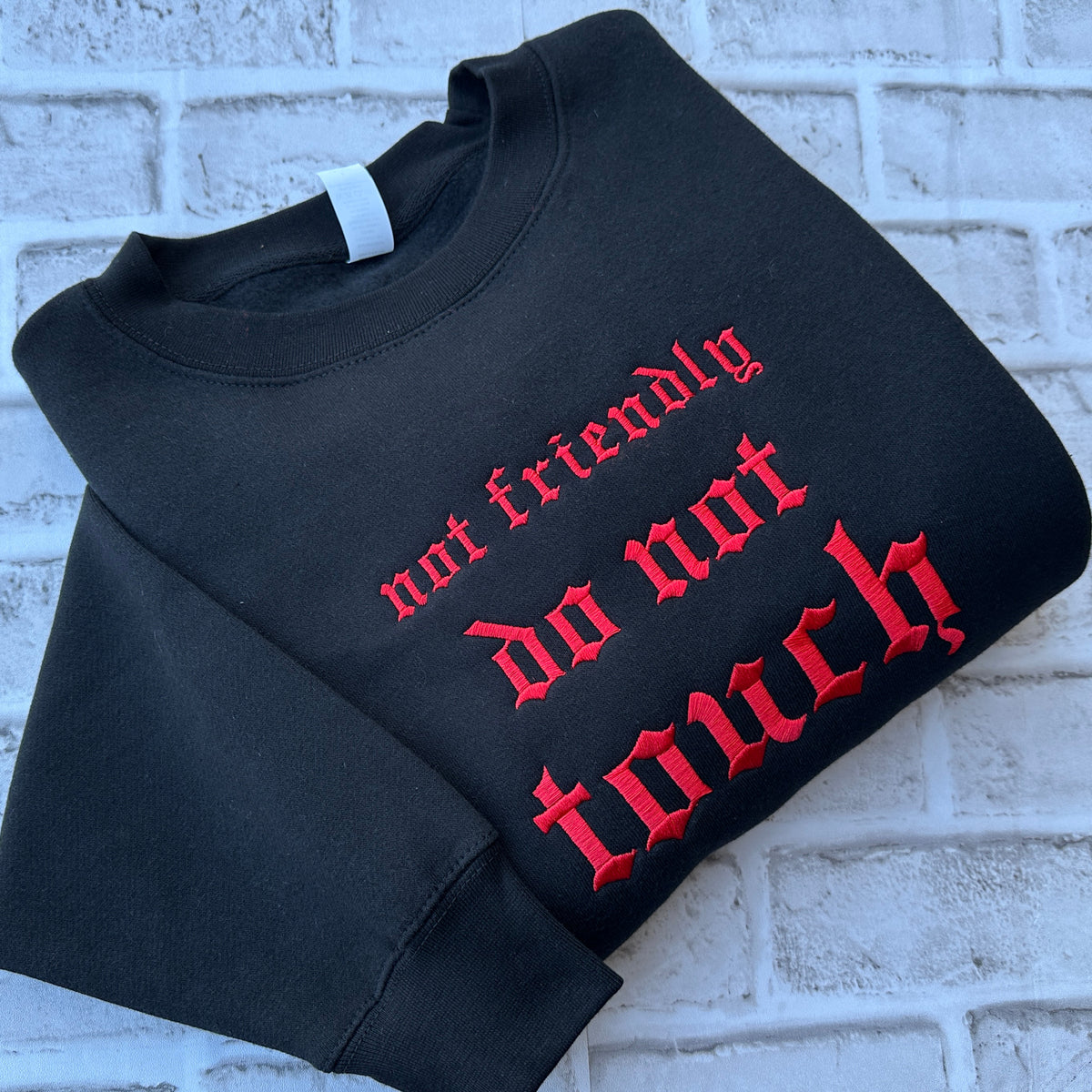 Black crewneck sweatshirt featuring bright red Old English embroidery that reads “not friendly do not touch” across the chest, with a classic round neckline, long sleeves, and ribbed cuffs and hem.