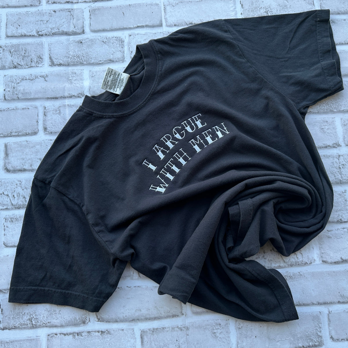 A black short-sleeve t-shirt with the phrase 'I ARGUE WITH MEN' embroidered in white arched text across the chest, laid flat on a white background.