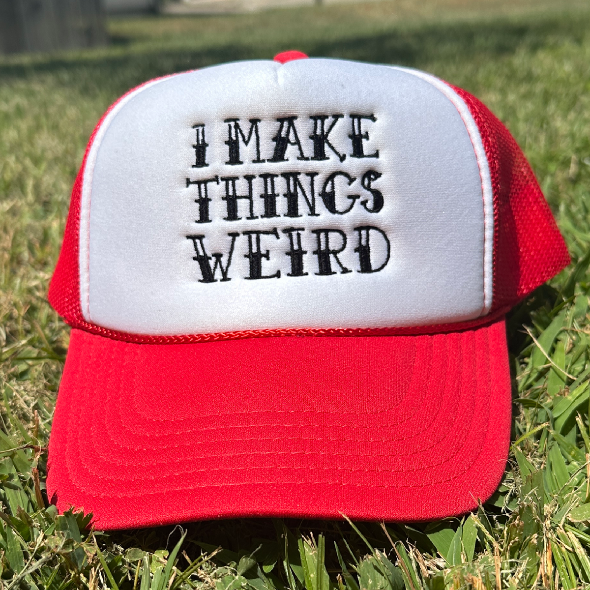 A red and white trucker hat with the embroidered text 'I MAKE THINGS WEIRD' on the front, resting on green grass outdoors.