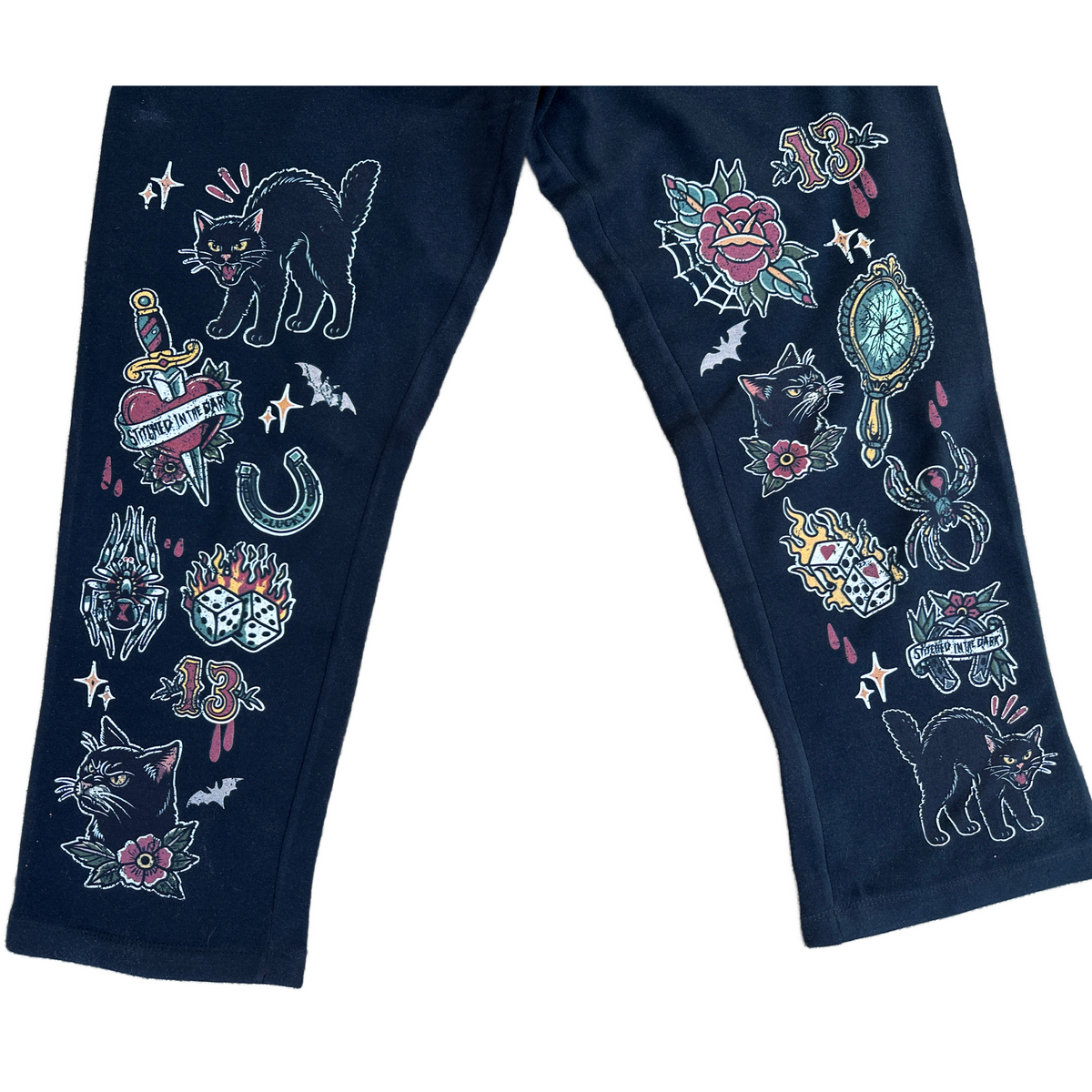 Black straight-leg sweatpants featuring colorful vintage tattoo-style graphics down both legs, including black cats, roses, bats, flaming dice, daggers, spiders, mirrors, horseshoes, number 13 motifs, and Stitched in the Dark banners.