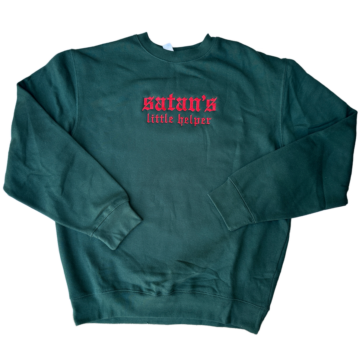 Dark forest green crewneck sweatshirt featuring red Old English gothic embroidery reading “satan’s little helper” across the upper chest. “Satan’s” appears in larger lettering above the smaller “little helper” text. Long-sleeve pullover with ribbed cuffs, collar, and hem, displayed flat against a white background.