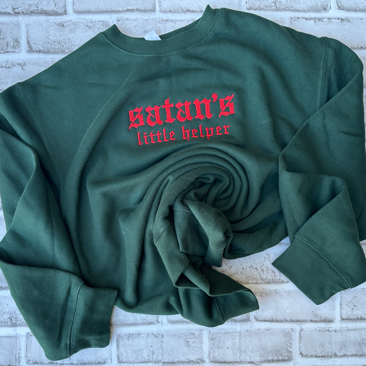 Dark forest green crewneck sweatshirt featuring red Old English gothic embroidery reading “satan’s little helper” across the upper chest. “Satan’s” appears in larger lettering above the smaller “little helper” text. Long-sleeve pullover with ribbed cuffs, collar, and hem, displayed flat against a white background.