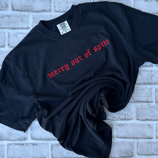 Black Comfort Colors short-sleeve T-shirt featuring red Old English gothic embroidery reading “merry out of spite” across the upper chest. Classic crewneck collar with a relaxed fit, displayed flat against a white background.