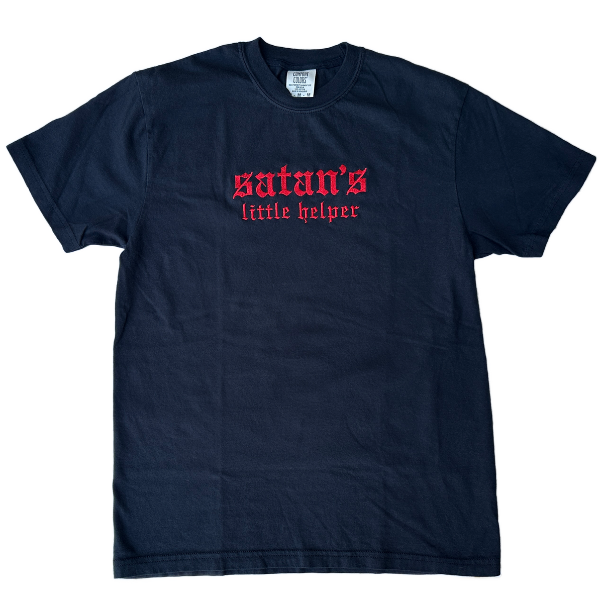 Black Comfort Colors short-sleeve T-shirt featuring red Old English gothic embroidery reading “satan’s little helper” across the upper chest. “Satan’s” appears in larger lettering above the smaller “little helper” text. Classic crewneck collar with a relaxed fit, displayed flat against a white background.