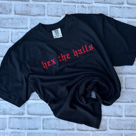 Black Comfort Colors short-sleeve T-shirt featuring red Old English gothic embroidery reading “hex the halls” across the upper chest. Classic crewneck collar with a relaxed fit, displayed flat against a white background.