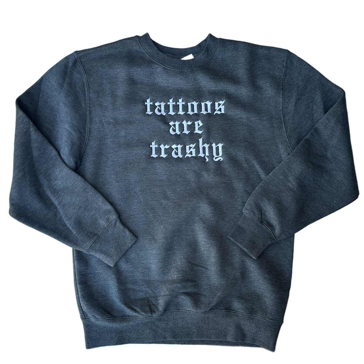 Dark heather gray crewneck sweatshirt featuring white embroidered “tattoos are trashy” lettering in lowercase Old English font, stacked across three lines on the chest.
