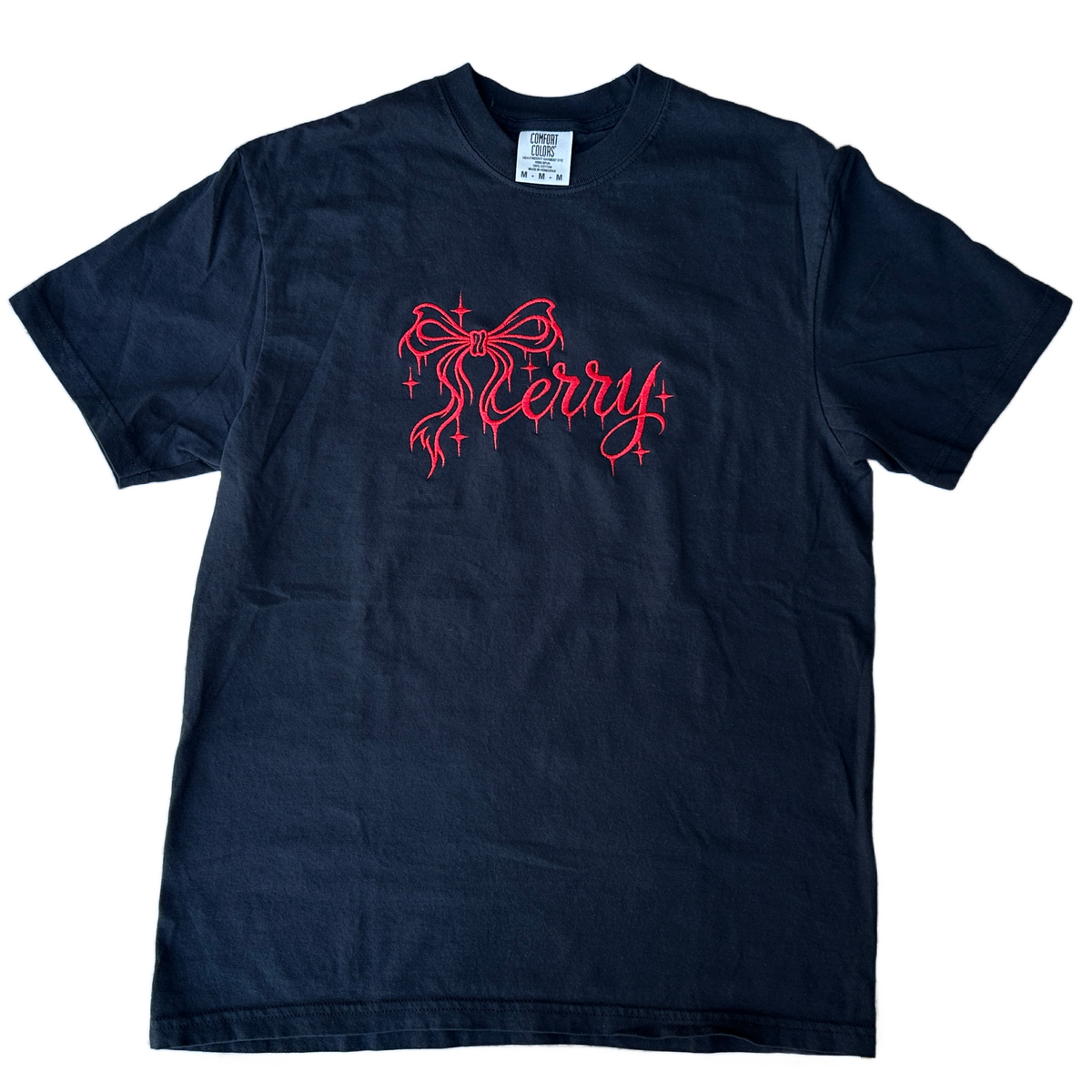 Black Comfort Colors T-shirt featuring bright pink embroidered “Merry” lettering in elegant script, accented with a decorative bow and sparkling stars across the chest.