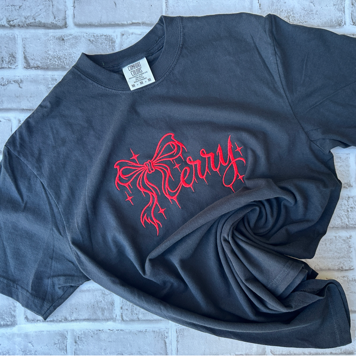 Black Comfort Colors T-shirt featuring bright pink embroidered “Merry” lettering in elegant script, accented with a decorative bow and sparkling stars across the chest.