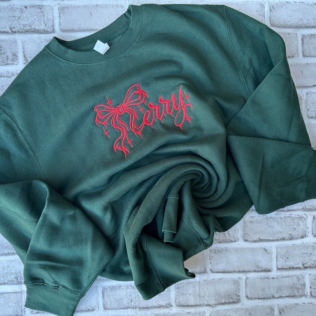 Dark forest green crewneck sweatshirt featuring red embroidered “Merry” lettering in elegant script, accented with a decorative bow and sparkling stars across the chest.