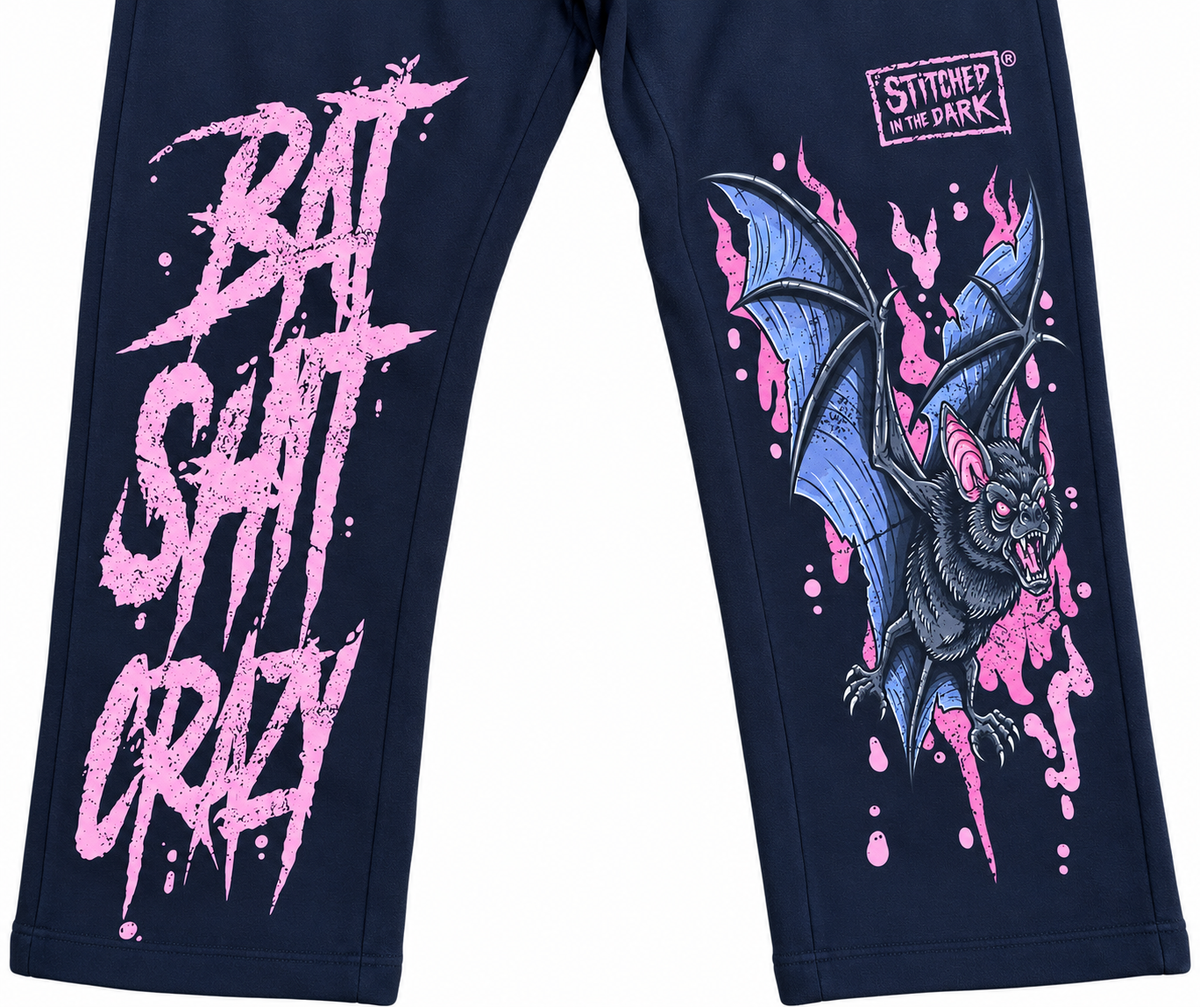Black sweatpants featuring a distressed pink “Bat Shit Crazy” graphic on one leg and a blue and black bat with pink accents and Stitched in the Dark logo on the other leg.
