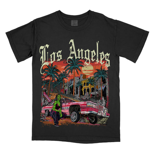 A black t-shirt  featuring the phrase ‘Los Angeles’ above a colorful illustration of palm trees, a lowrider car, and a sunset city scene. Zombie girl models by car.