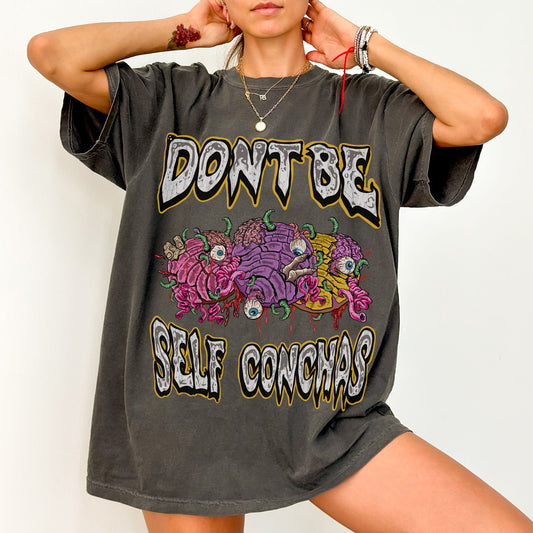 Model wearing an oversized pepper color comfort colors graphic T-shirt featuring colorful illustrated spooky conchas and text reading “Don’t Be Self Conchas.”