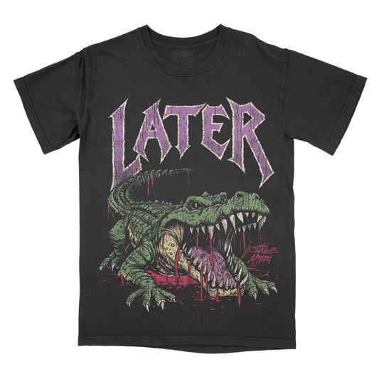 A black short-sleeve graphic t-shirt featuring a green alligator illustration with its mouth open, dripping with purple liquid, beneath the word 'LATER' in a stylized purple font.