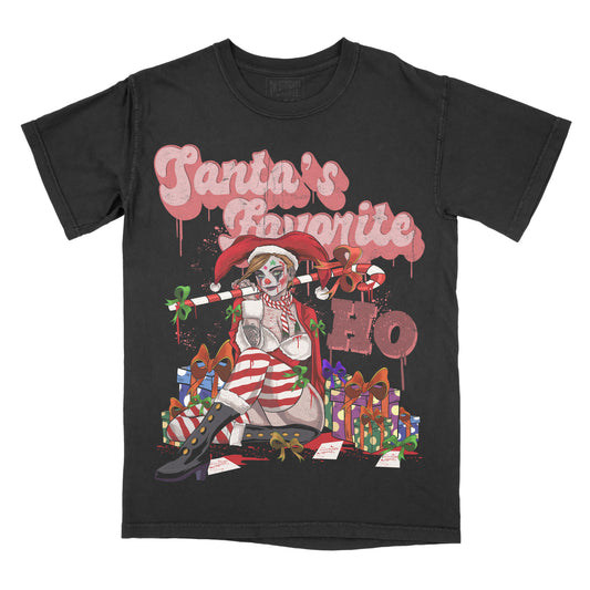 Black graphic t-shirt featuring the phrase “Santa’s Favorite Ho” in dripping pink lettering above an illustrated gothic Santa-style character surrounded by wrapped gifts and holiday-themed details.