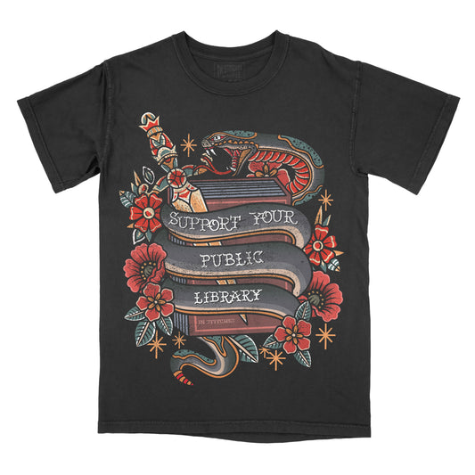 A black short-sleeve crewneck t-shirt featuring a detailed graphic print on the front. The graphic depicts a traditional tattoo-style design with a snake wrapped around a book, red flowers, and banners reading 'SUPPORT YOUR PUBLIC LIBRARY' 